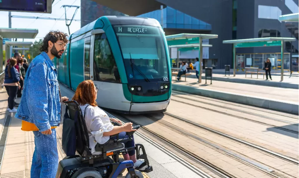 A young man accompanied a woman in a wheelchair as they waited for a modern tram to arrive at the station.
