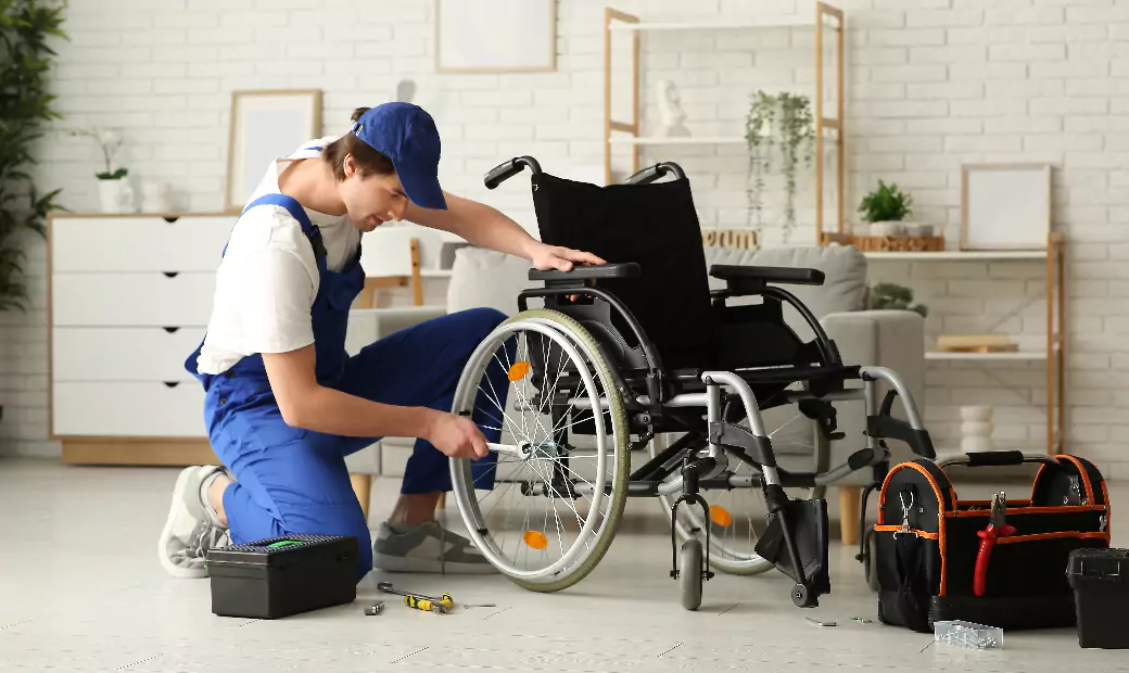 Technician in blue uniform repairing a manual wheelchair indoors with toolboxes nearby.