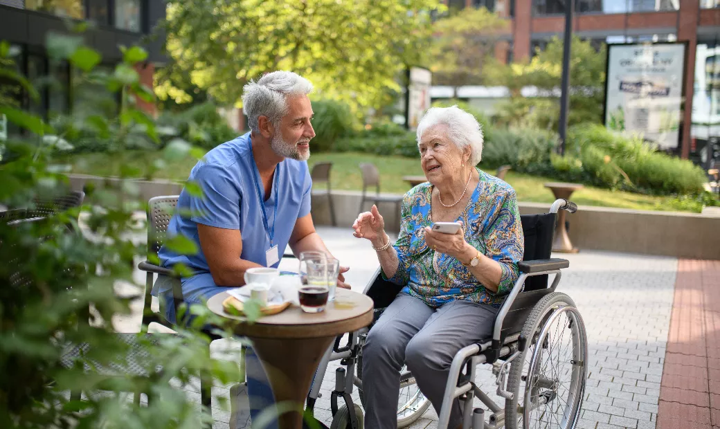 a-male-nurse-is-sitting-in-a-wheelchair-with-an-elderly-patient-drinking-coffee-together